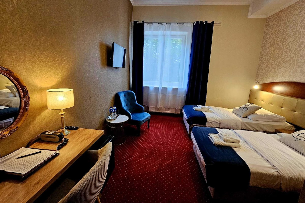 A hotel room with two beds, a desk, a chair, a TV, and a window with curtains.