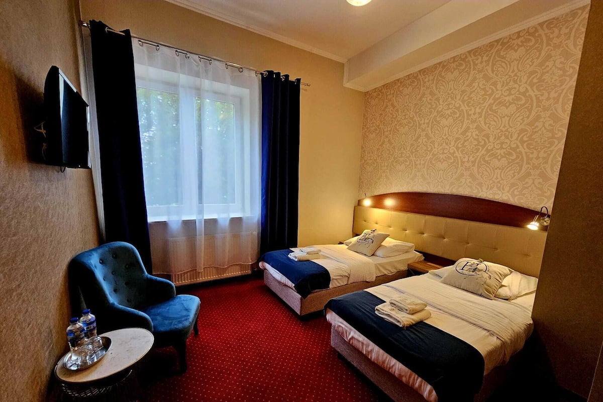 A hotel room with two beds, a chair, a small table with water bottles, and a TV on the wall.