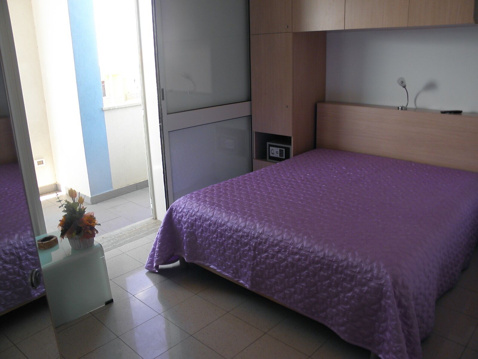 A hotel room with a bed, a small table with a vase of flowers, a desk with a computer, and a window with a view of the outside.