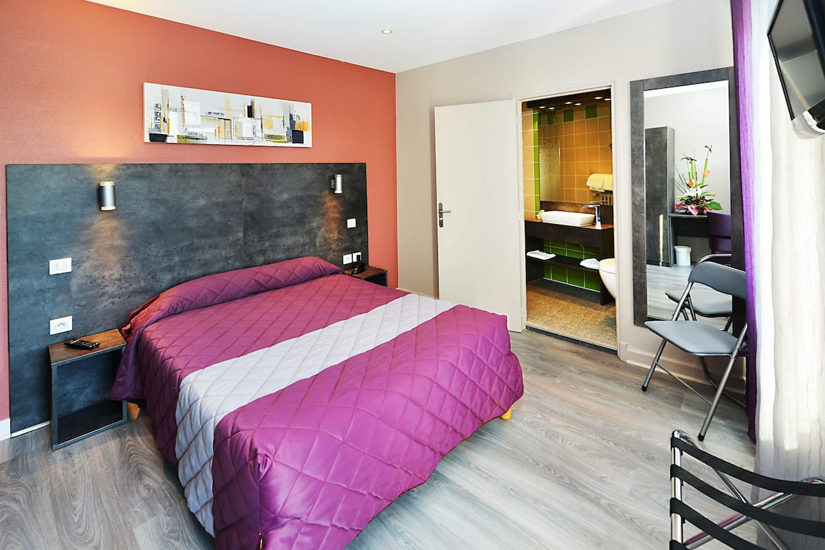 A modern hotel room with a large bed, a nightstand, a desk with a chair, and a bathroom visible through an open door.