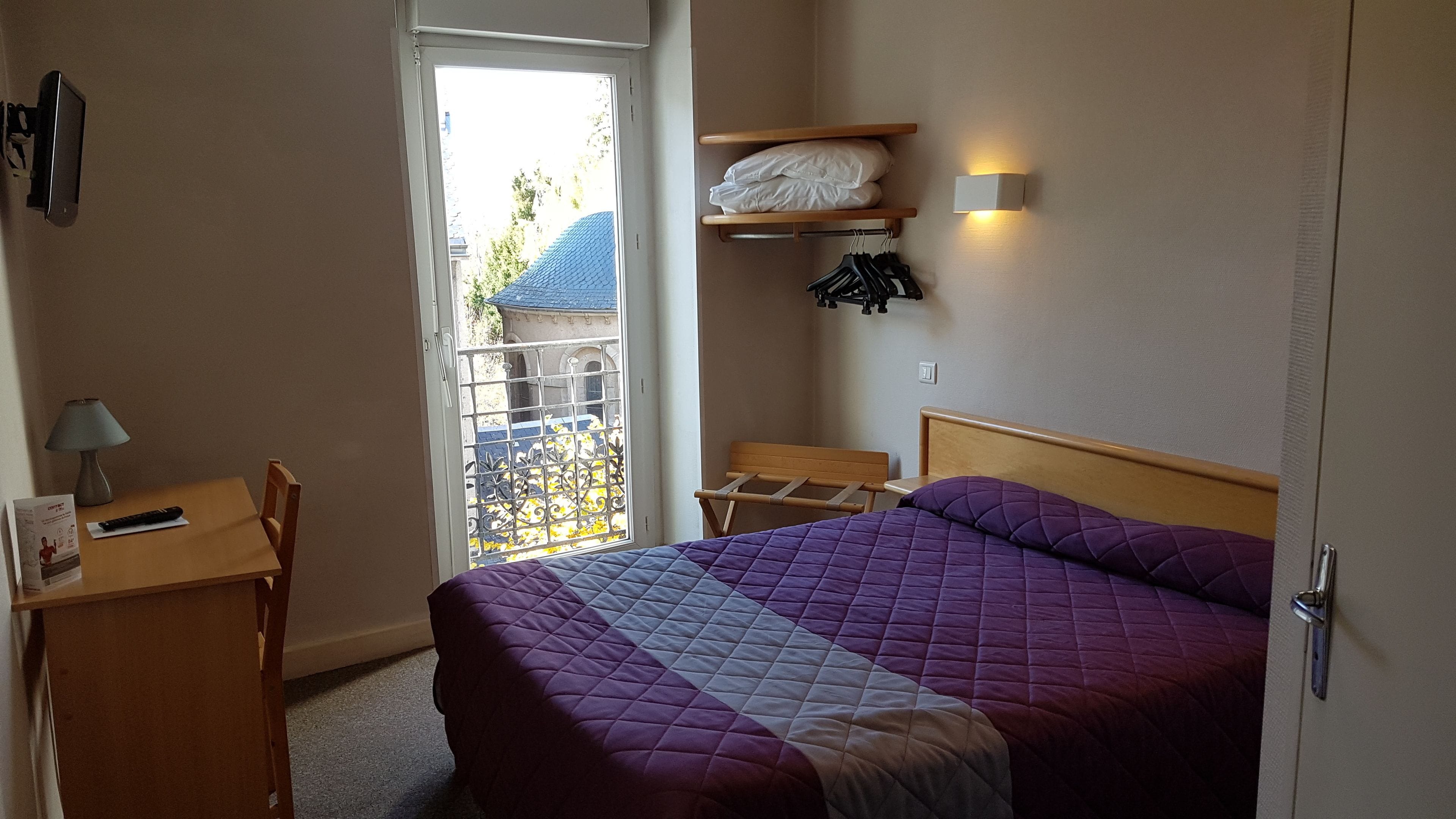 Habitación de hotel con cama, escritorio, silla, lámpara, armario y ventana con vistas al exterior.