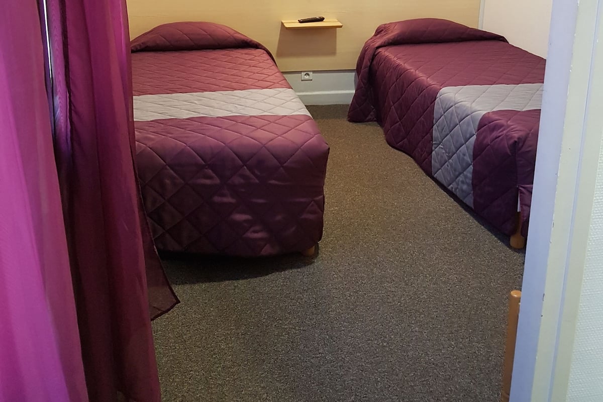 A room with two beds, a nightstand, and a wall-mounted light.