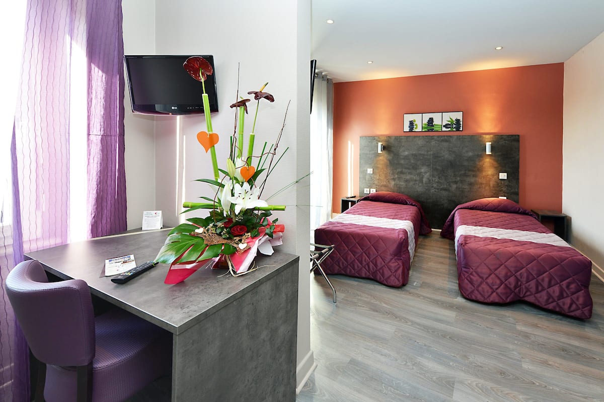 A hotel room with a desk, two beds, a TV, and a flower arrangement.
