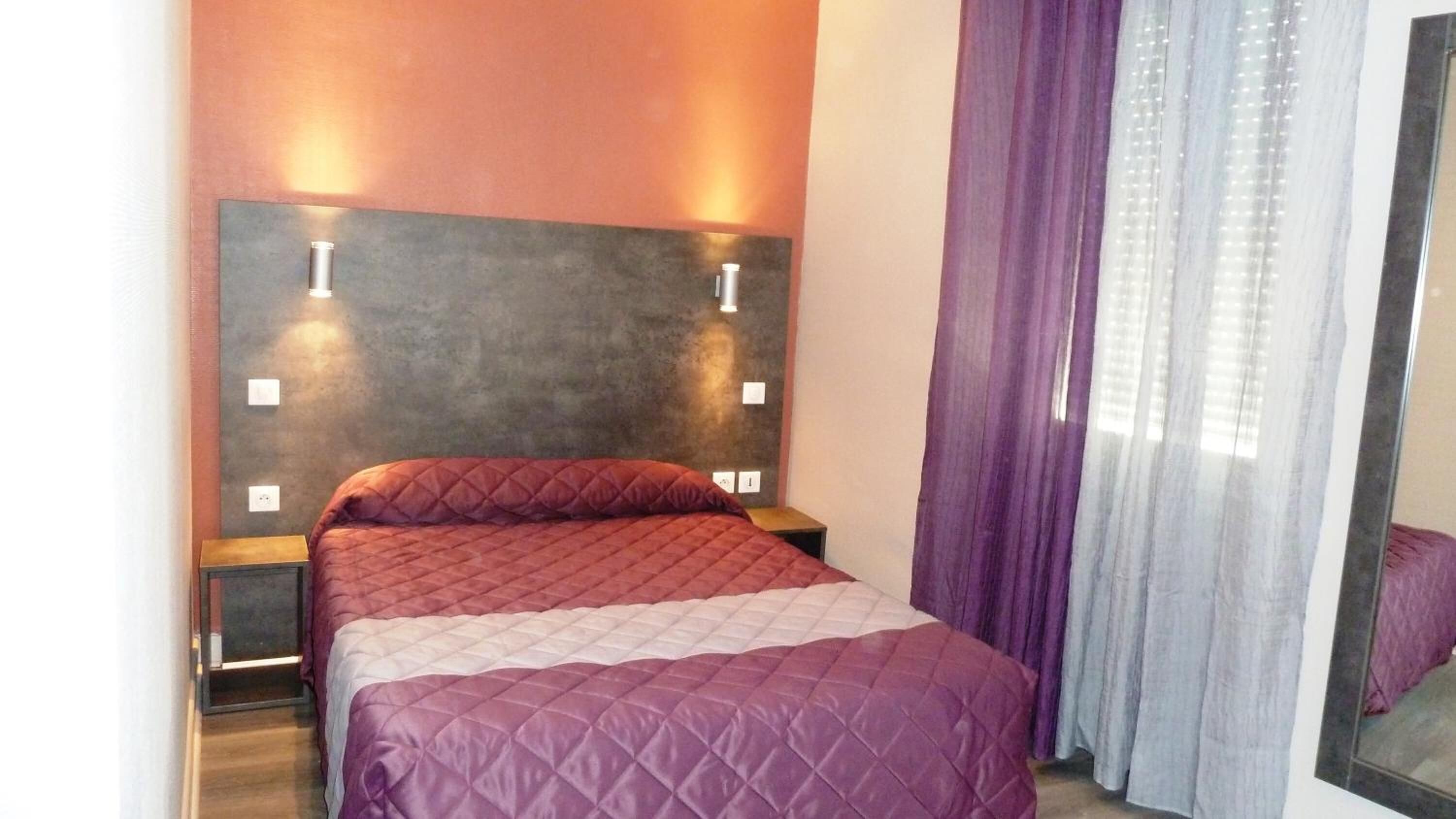 Una habitación de hotel con cama, cabecero con luces fijadas a la pared, mesita de noche y ventana con cortinas moradas.