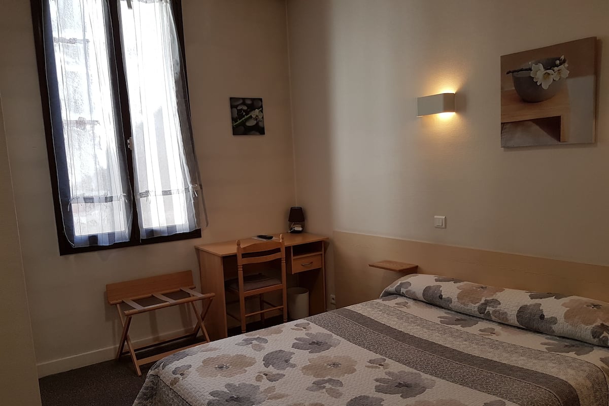 A bedroom with a bed, a desk, and a chair. There is a window with curtains, a wall-mounted light, and a small shelf.