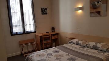 Un dormitorio con cama, escritorio y silla. Se observa una ventana con cortinas, una lámpara de pared y un estante pequeño.