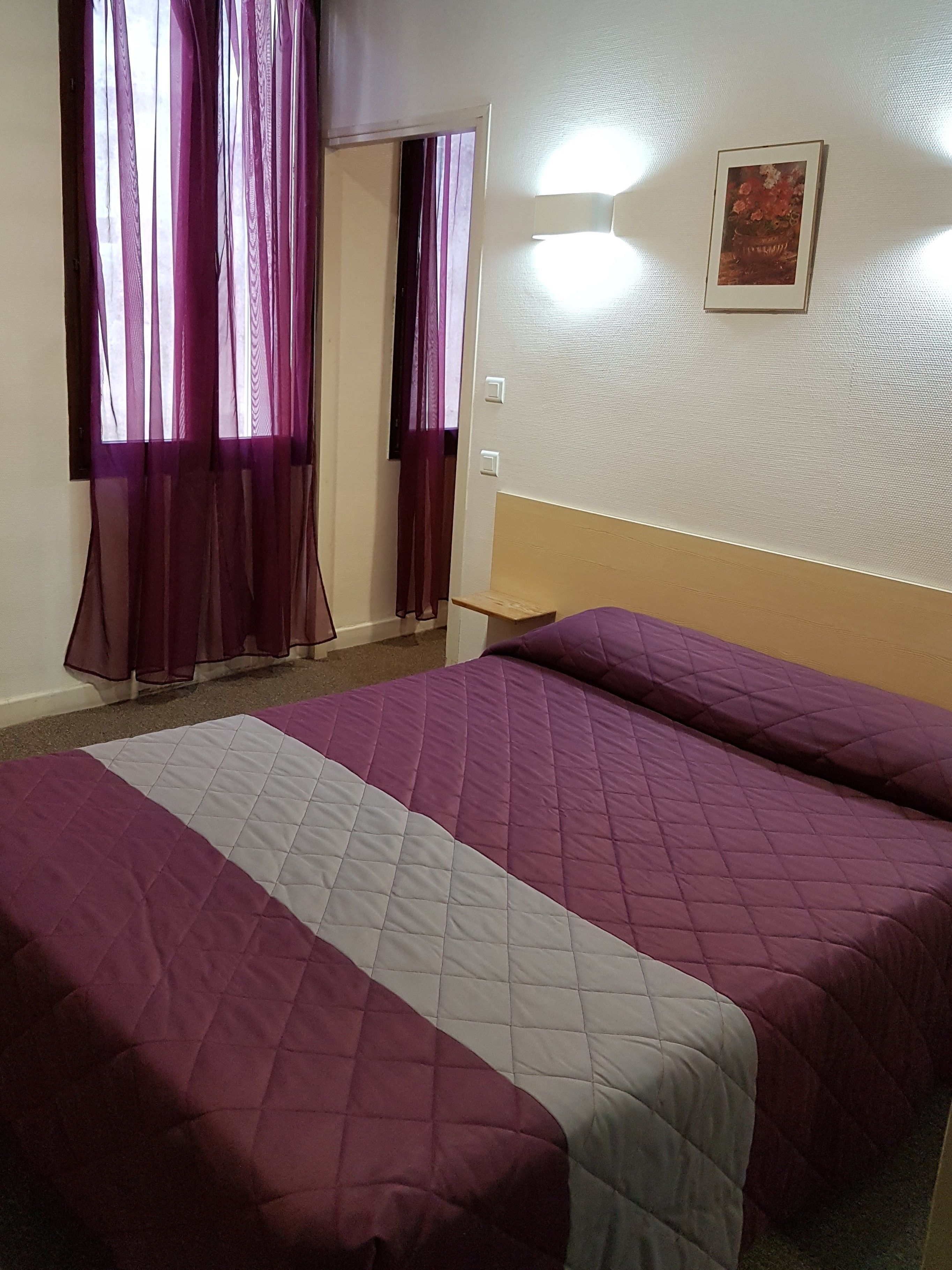 Habitación de hotel con una cama grande, cortinas moradas, una mesita de noche pequeña y un cuadro enmarcado en la pared.