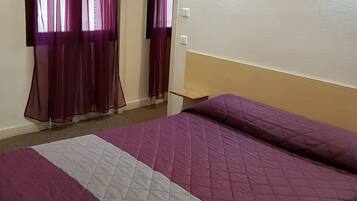 Habitación de hotel con una cama grande, cortinas moradas, una mesita de noche pequeña y un cuadro enmarcado en la pared.