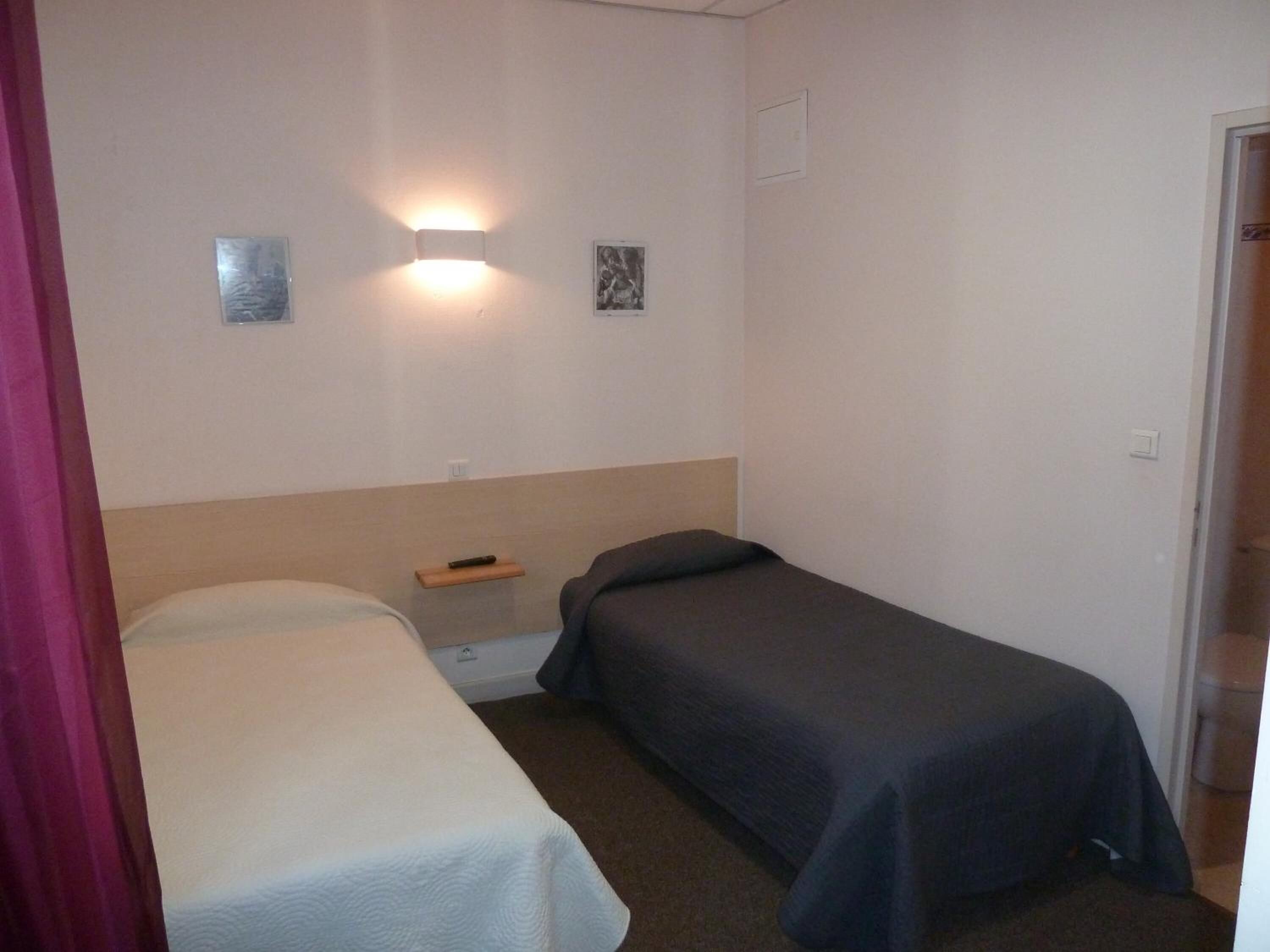 A small, simple room with two single beds, a nightstand, and a wall-mounted light fixture.