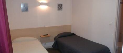 A small, simple room with two single beds, a nightstand, and a wall-mounted light fixture.