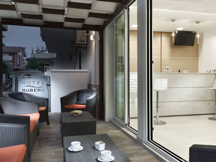 A hotel lobby with a modern design, featuring a reception desk, seating area with sofas and tables, and a large glass door leading to the outside.