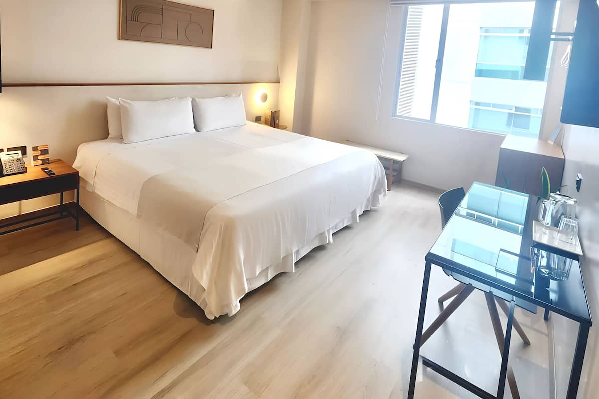 A modern hotel room with a large bed, bedside tables, a desk, and a window with blinds.