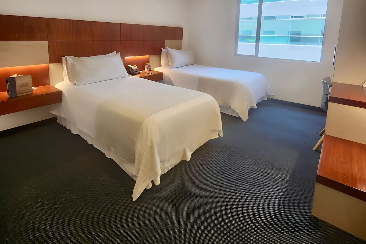 A hotel room with two beds, a wooden headboard, a bedside table with a phone, a nightstand, a window with a view of buildings, and a desk with a chair.