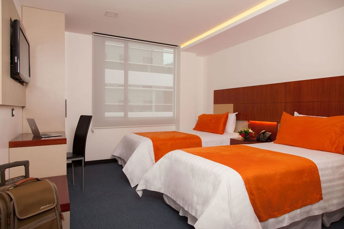 A hotel room with two beds, a desk with a laptop, a chair, a suitcase, and a window with blinds.