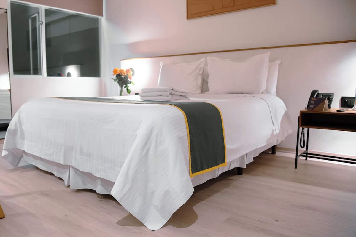 A neatly made bed with a white and green bedspread, a bedside table with a phone and a small plant, and a chair with a wooden frame.