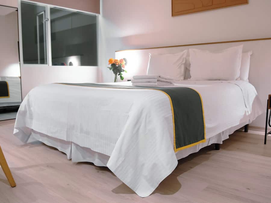 A neatly made bed with a white and green bedspread, a bedside table with a phone and a small plant, and a chair with a wooden frame.