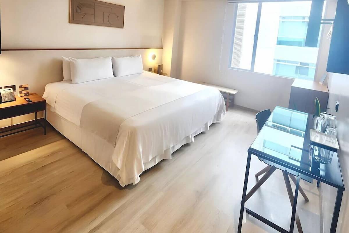 A modern hotel room with a large bed, bedside table, lamp, and a desk with a chair.