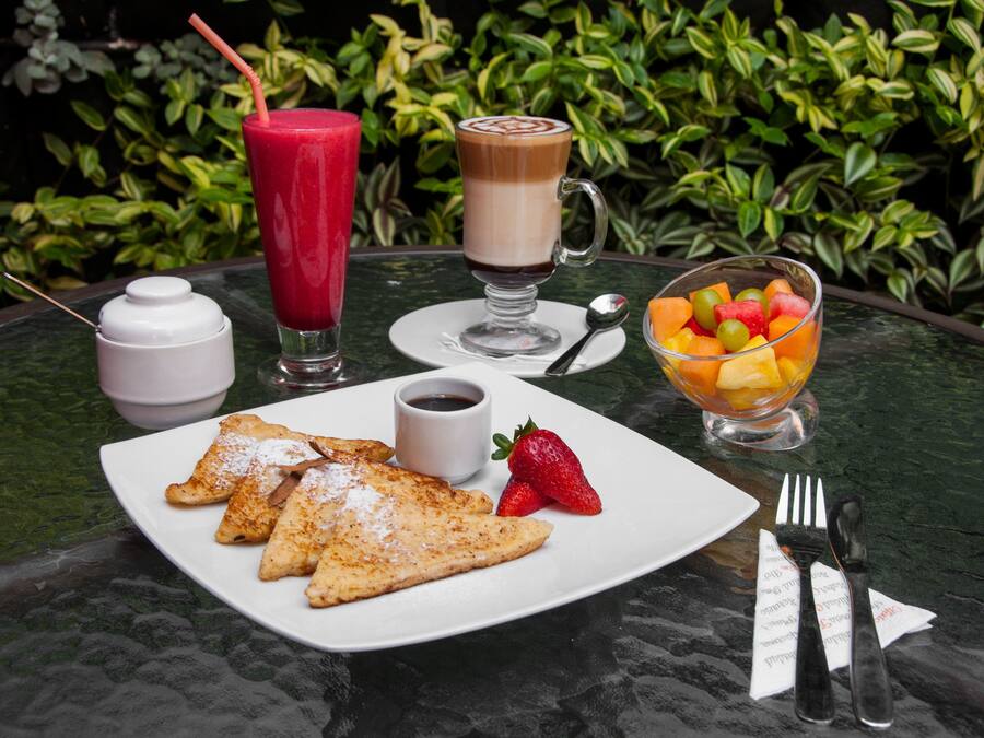 Una mesa con alimentos para el desayuno, incluyendo panqueques, un bol de fruta, una bebida de café y un batido.