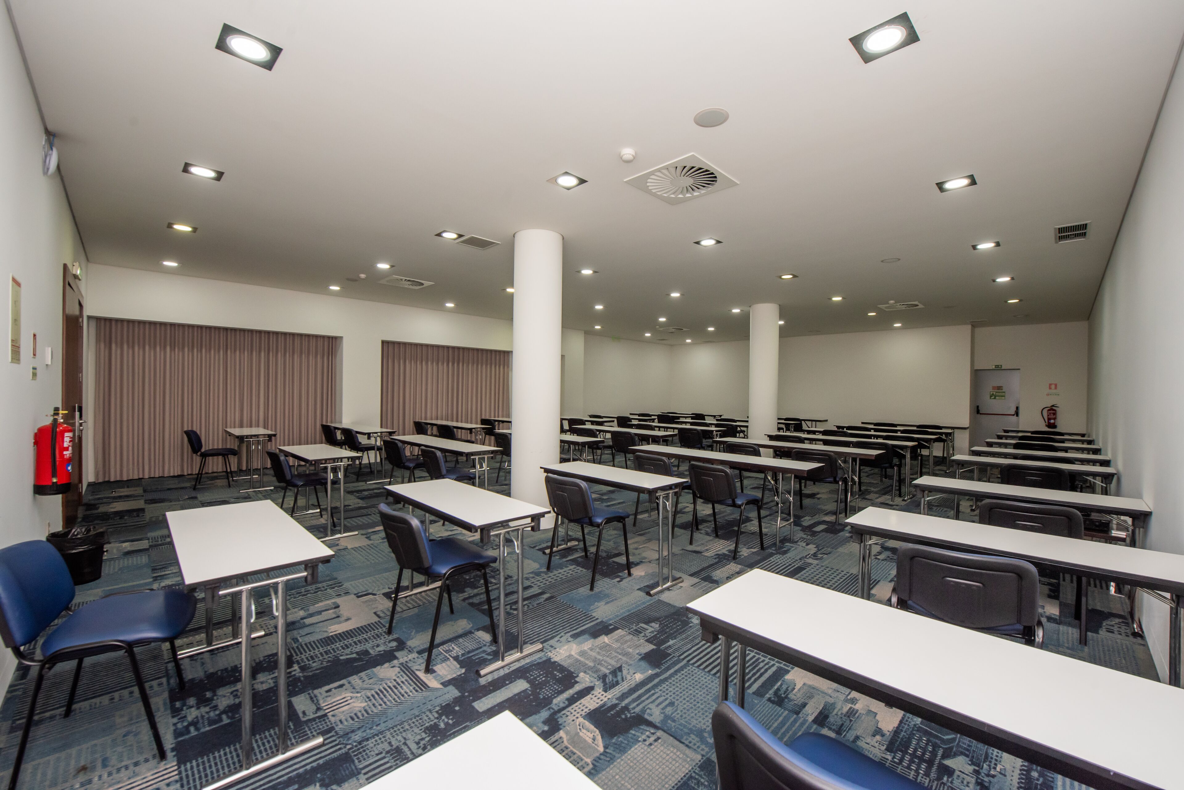 Un salón de conferencias con varias mesas y sillas, un extintor y una alfombra estampada.