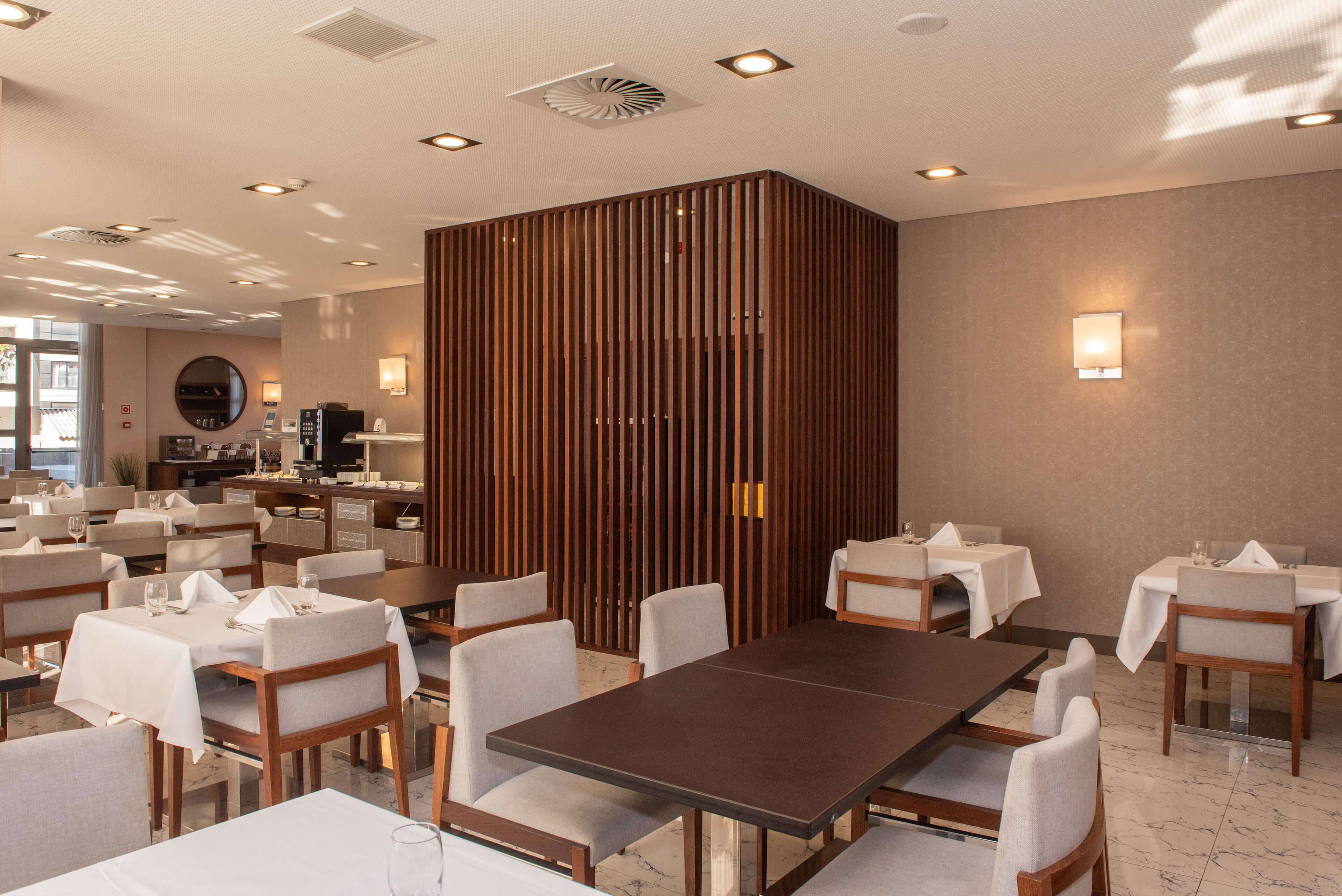Un comedor moderno con varias mesas y sillas, un separador central de madera y una cocina al fondo.