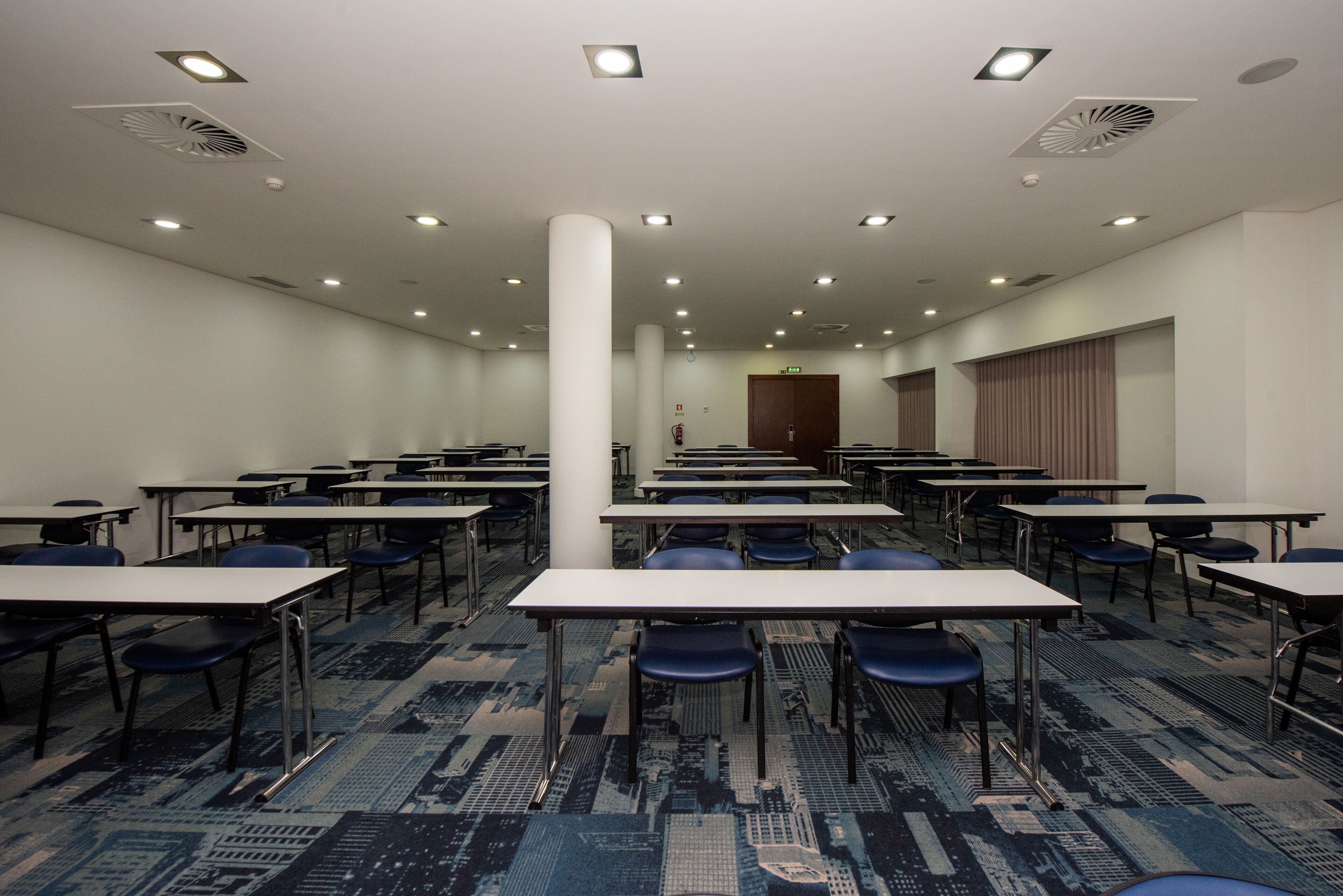 A conference room with rows of tables and chairs, white walls, and a patterned carpet.