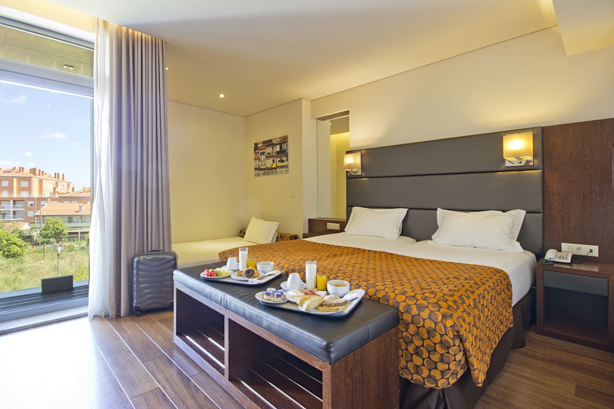 A hotel room with a bed, a bench with breakfast trays, a suitcase, and a view of buildings outside.