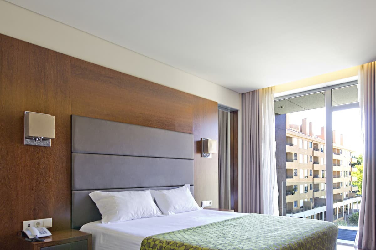 A hotel room with a large bed, a wooden headboard, a bedside table with a phone, and a view of a building through the window.