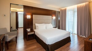 A modern hotel room with a large bed, wooden flooring, a desk with a chair, a nightstand, and a large window with curtains.
