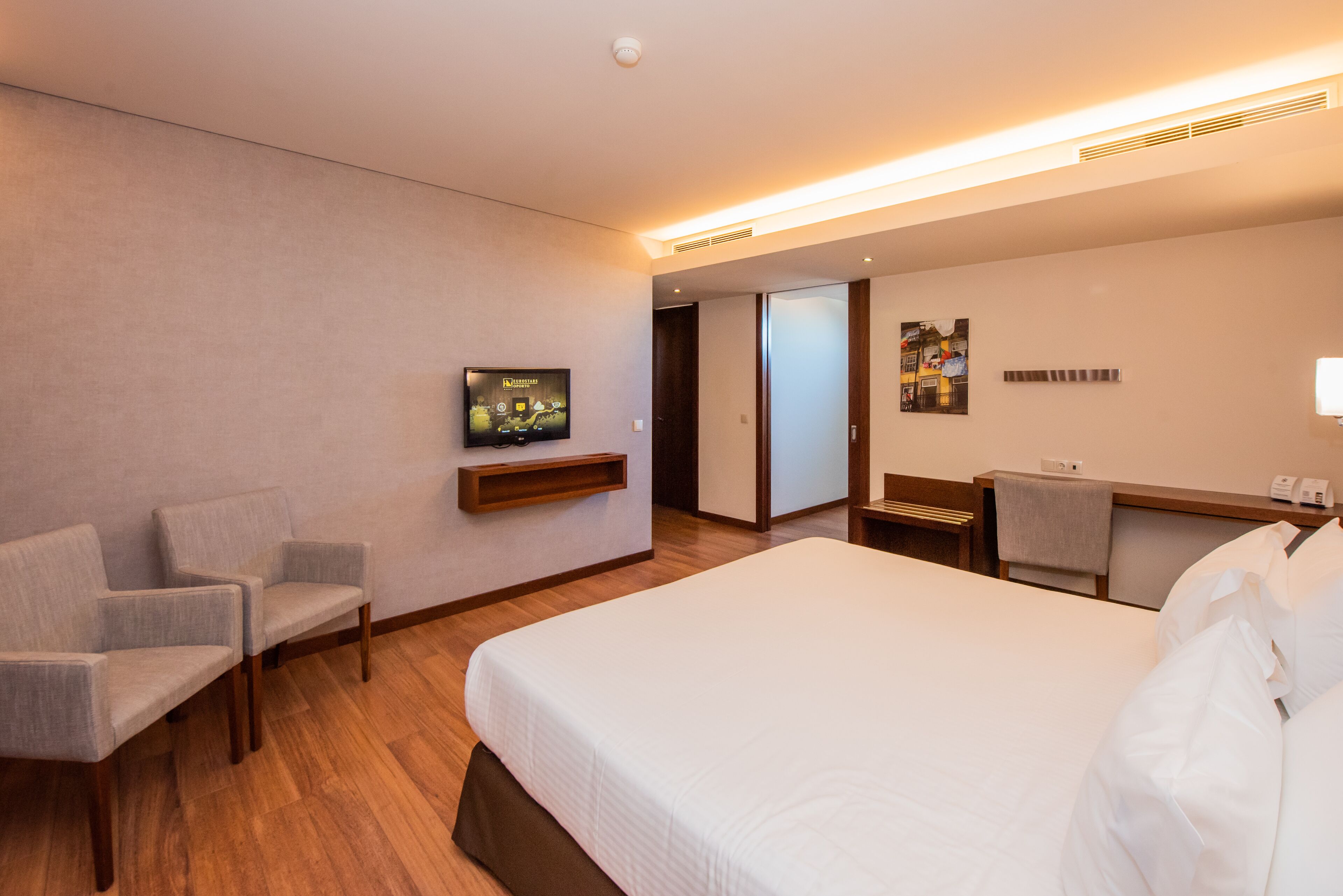 Quarto de hotel com uma cama grande, duas poltronas, uma mesa de trabalho com cadeira, televisão e uma porta que dá para outro cômodo.