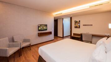 Quarto de hotel com uma cama grande, duas poltronas, uma mesa de trabalho com cadeira, televisão e uma porta que dá para outro cômodo.