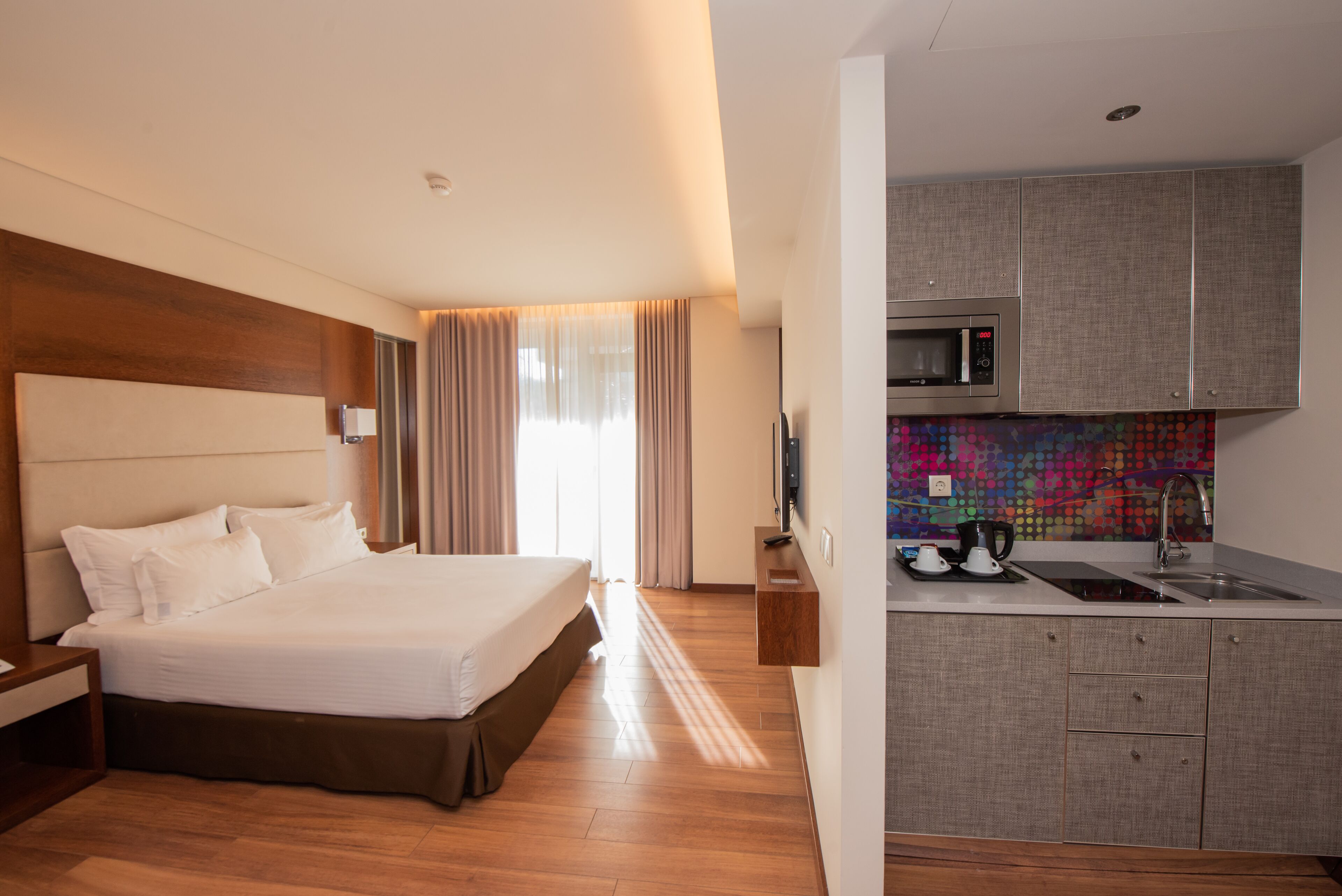 Una camera d'albergo con un letto grande, una testiera in legno, un comodino, una TV integrata e un angolo cottura dotato di microonde, lavello e zoccolo colorato.