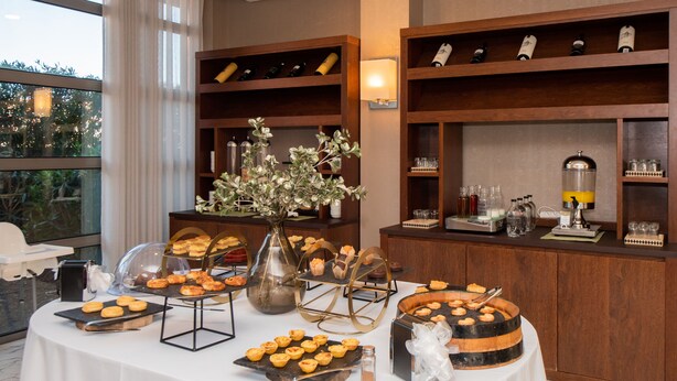 A buffet table with various pastries and drinks, surrounded by wooden shelves with wine bottles.