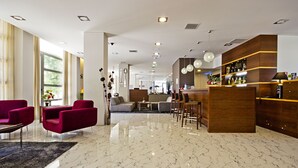 A modern hotel lobby with a marble floor, red armchairs, a bar area, and a seating area with sofas and a coffee table.