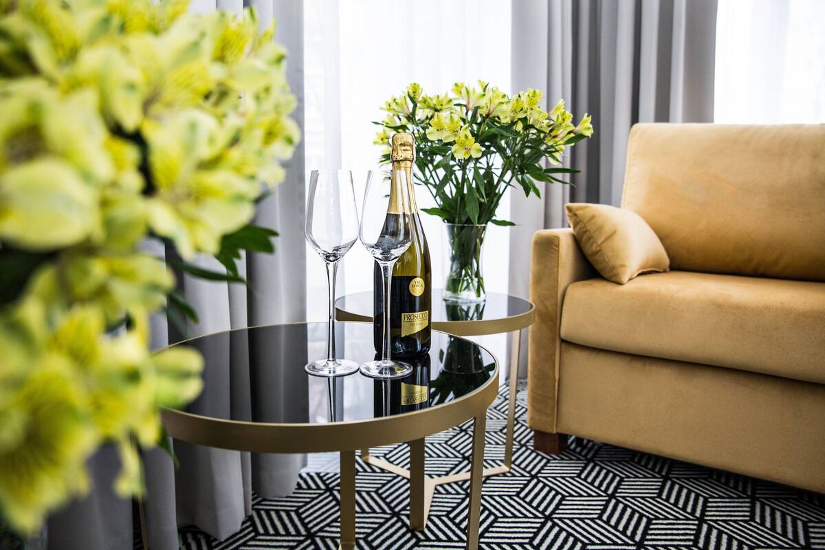 A round glass table with a bottle of champagne and two wine glasses, a vase with yellow flowers, and a beige sofa in a modern room with patterned flooring.