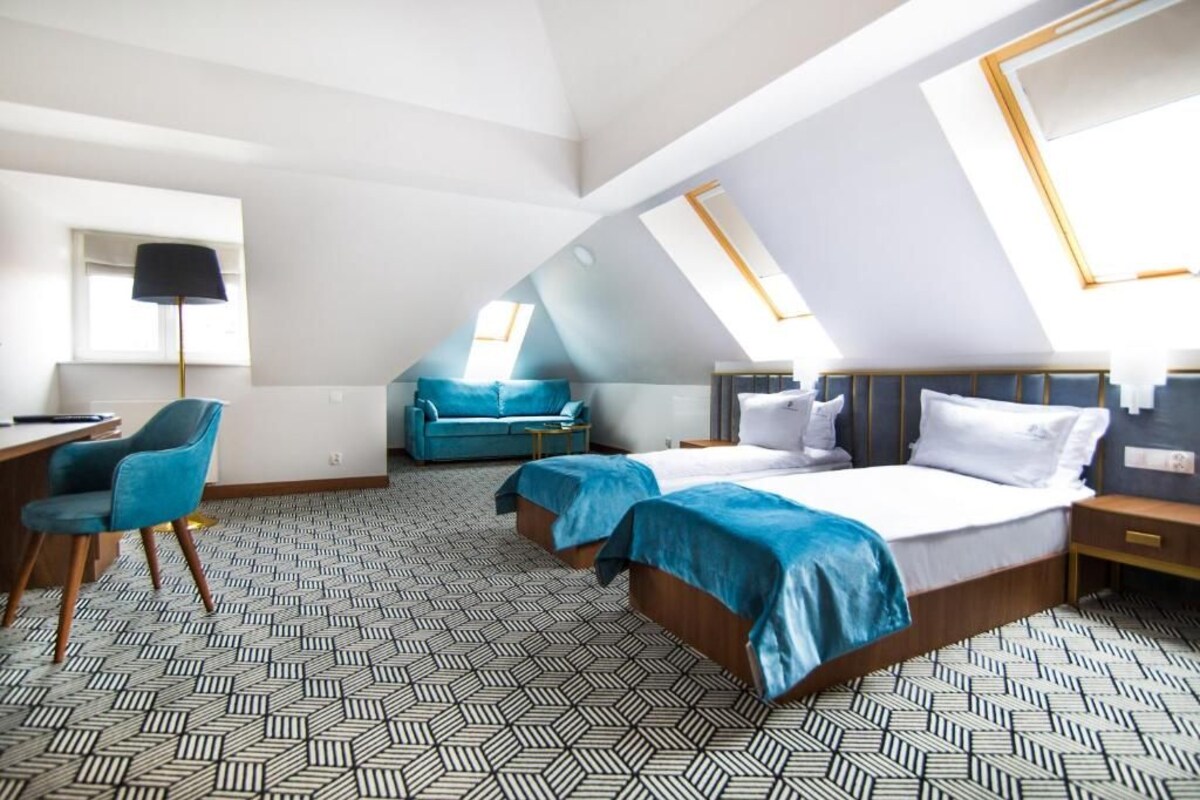 A modern hotel room with a sloped ceiling, two beds with blue bedding, a desk, and a chair.