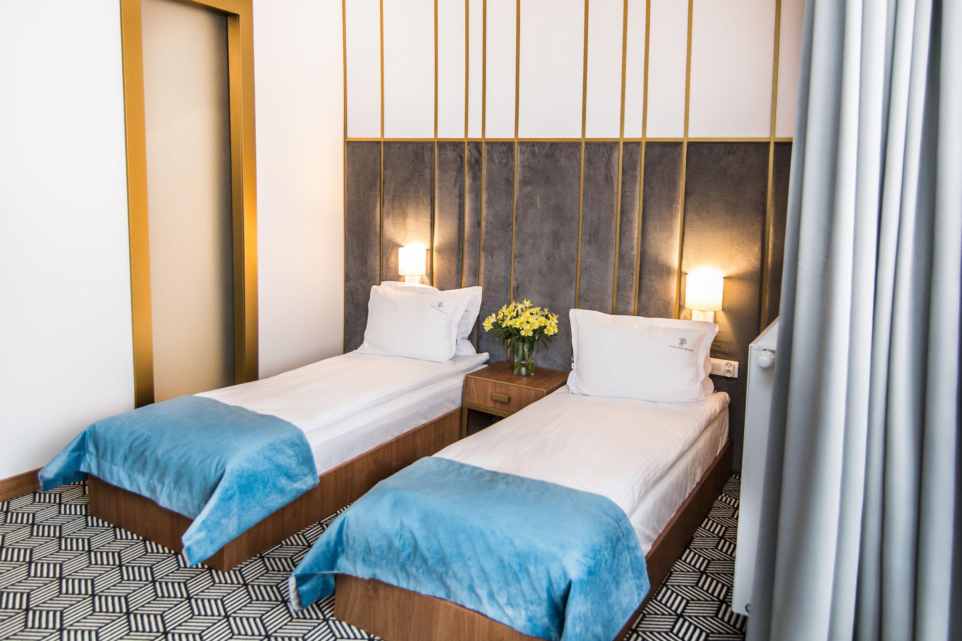 Een hotelkamer met twee eenpersoonsbedden, een nachtkastje met een lamp, een bloemenvaas en een vloer met een patroon.