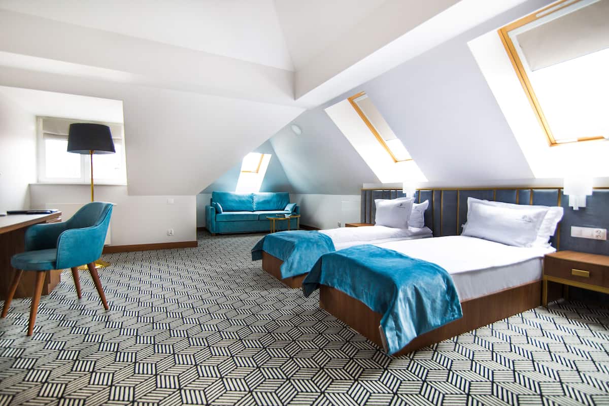 A modern hotel room with a geometric patterned floor, a desk with a chair, two beds with blue bedding, and a sofa.