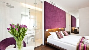 A modern hotel room with a large bed, a glass shower enclosure, and a purple accent wall.