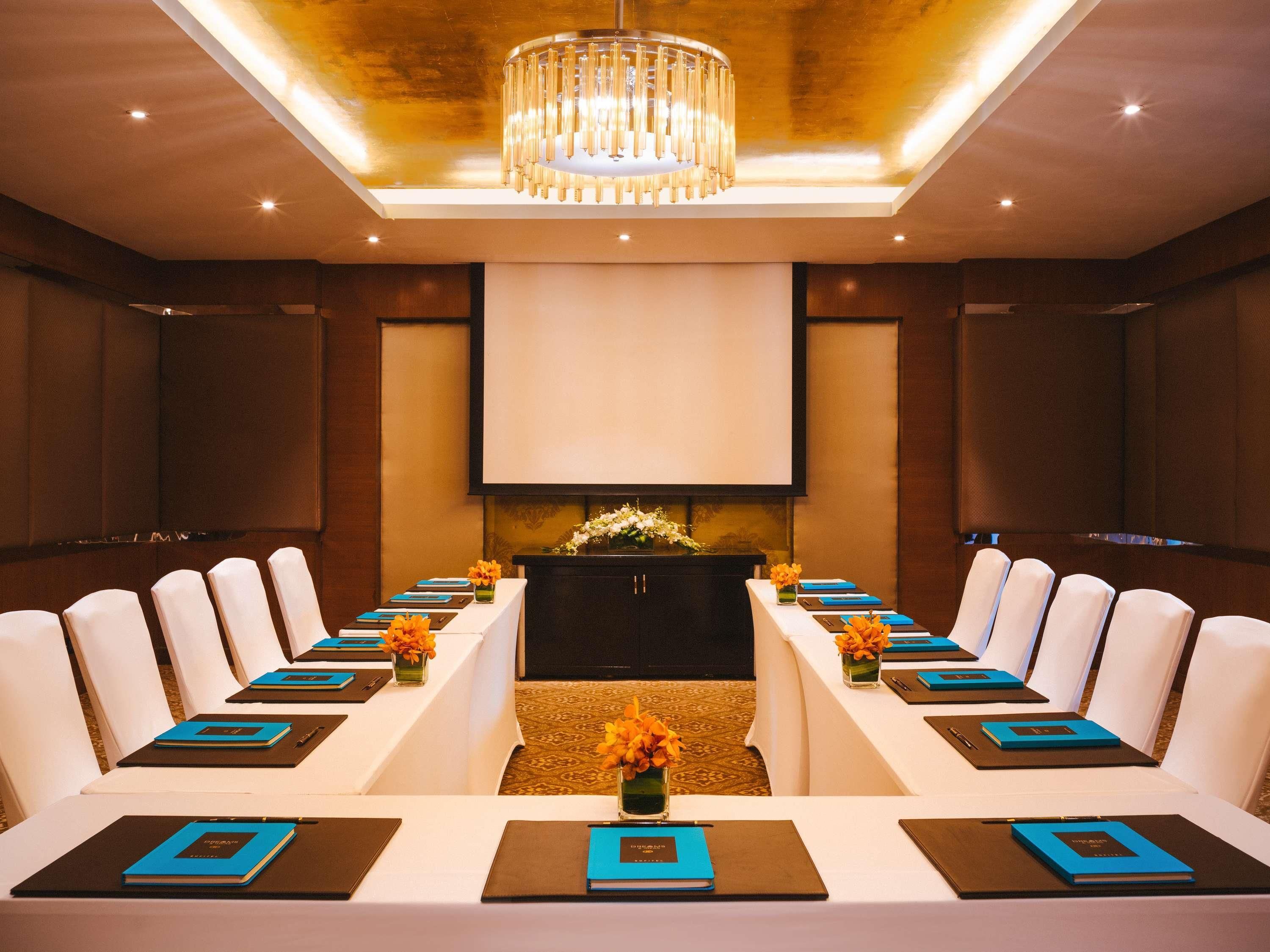 Uma sala de conferências com uma longa mesa, cadeiras brancas, uma tela de projetor e um candelabro.
