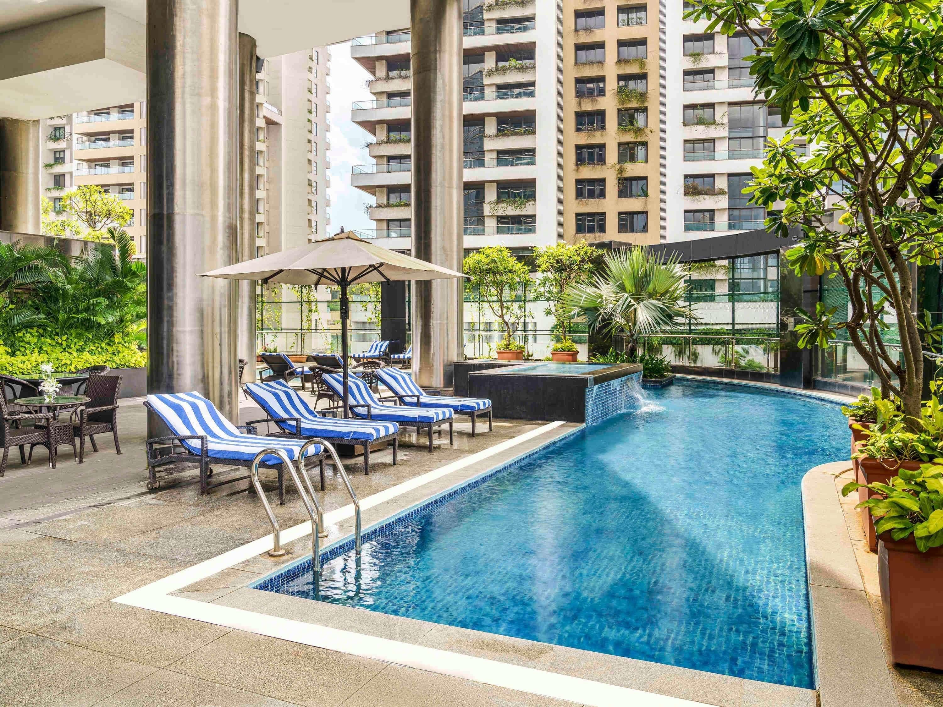 A swimming pool with lounge chairs and umbrellas, surrounded by greenery and tall buildings.