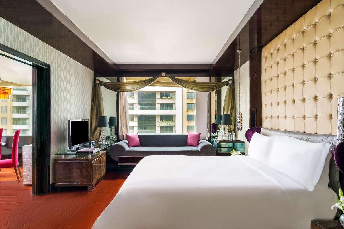 A hotel room with a large bed, a desk, a sofa, and a view of buildings through the window.