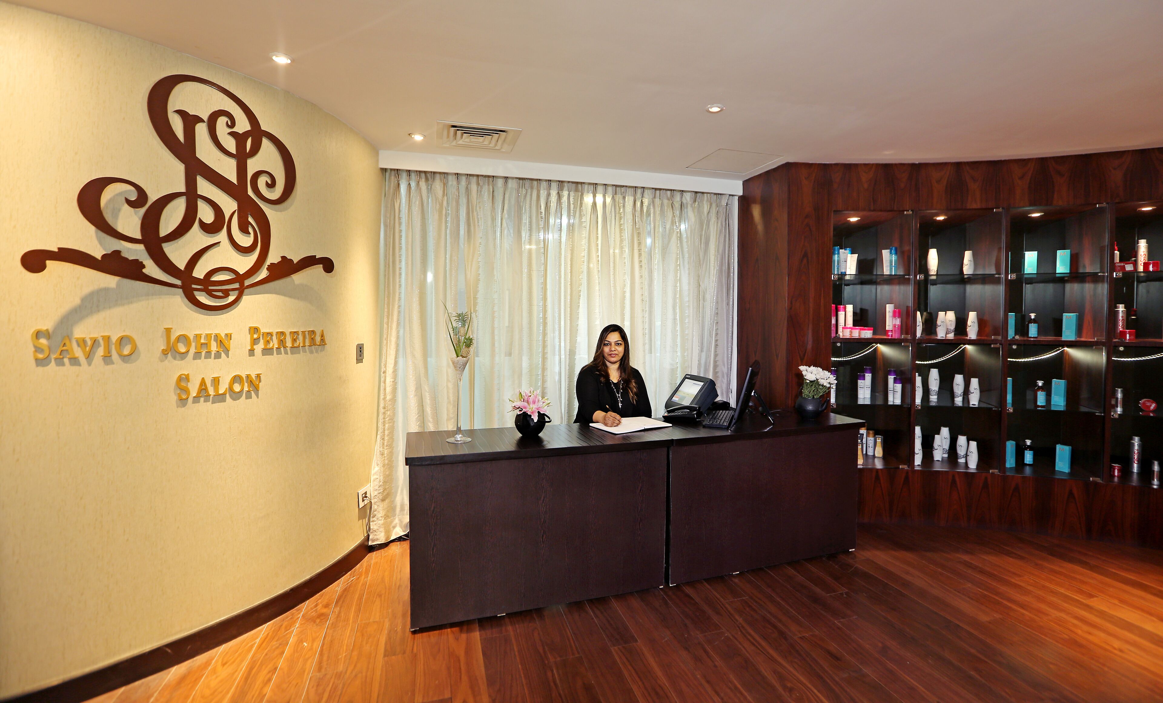 A reception desk with a woman seated behind it, a wall sign reading "Savio John Pereira Salon", and a shelf with various products.