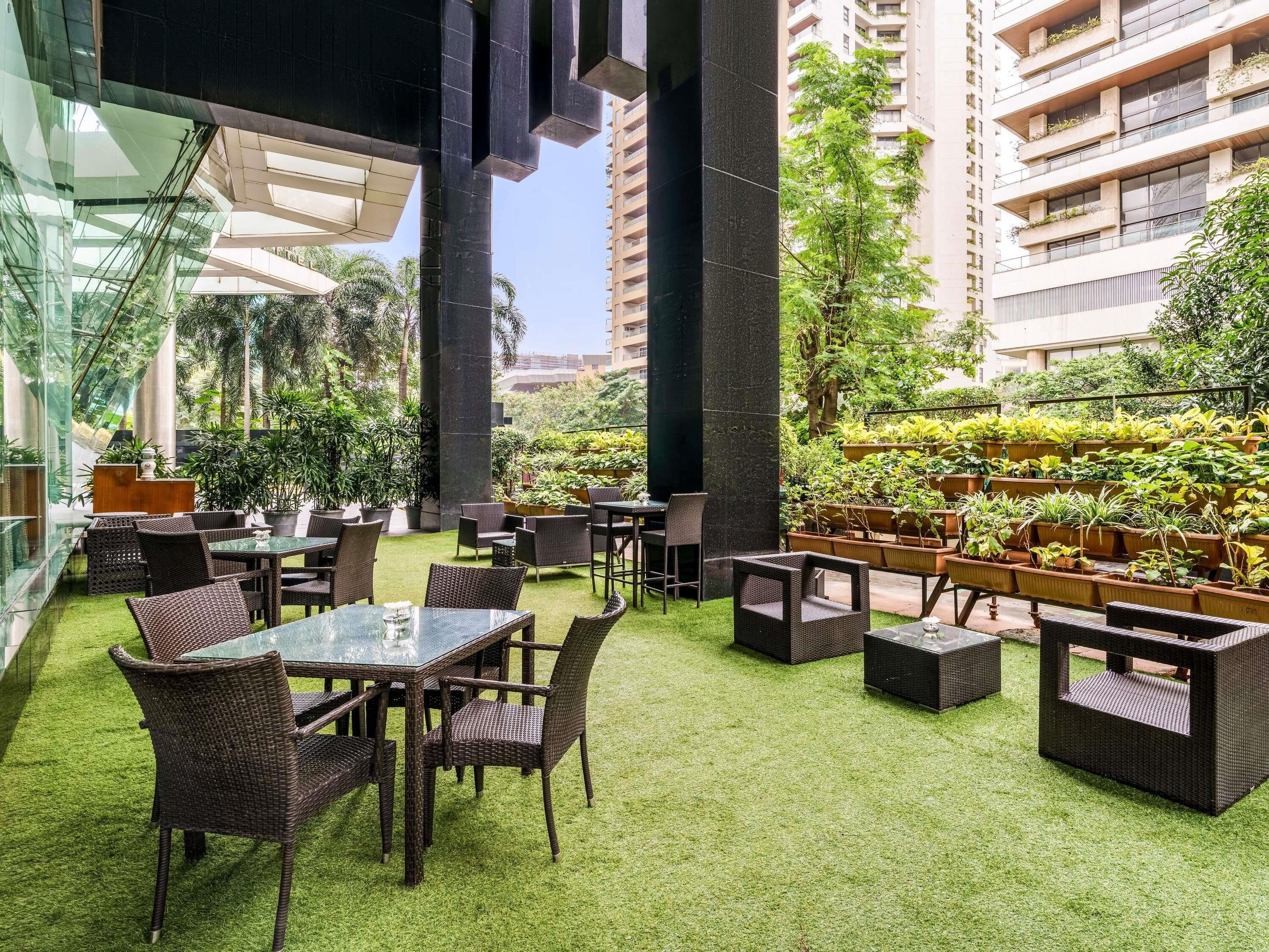 Een terras met tafels en stoelen, omringd door groen en hoge gebouwen.