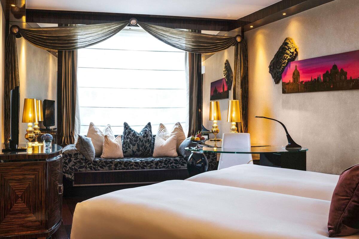 A hotel room with a large bed, a desk with two lamps, a sofa with cushions, a nightstand, and a painting on the wall.