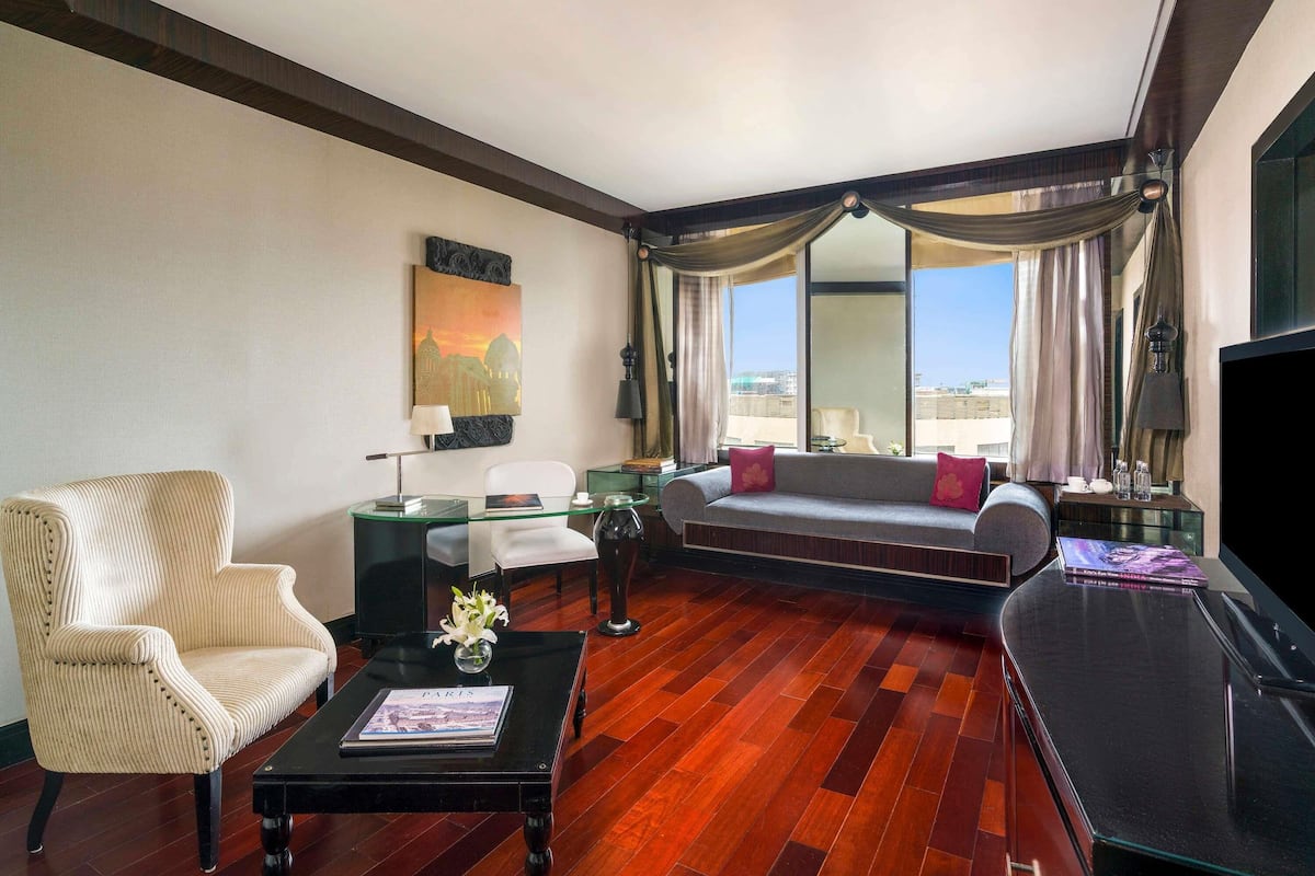 A hotel room with a sofa, armchair, coffee table, TV, and a large window with curtains.