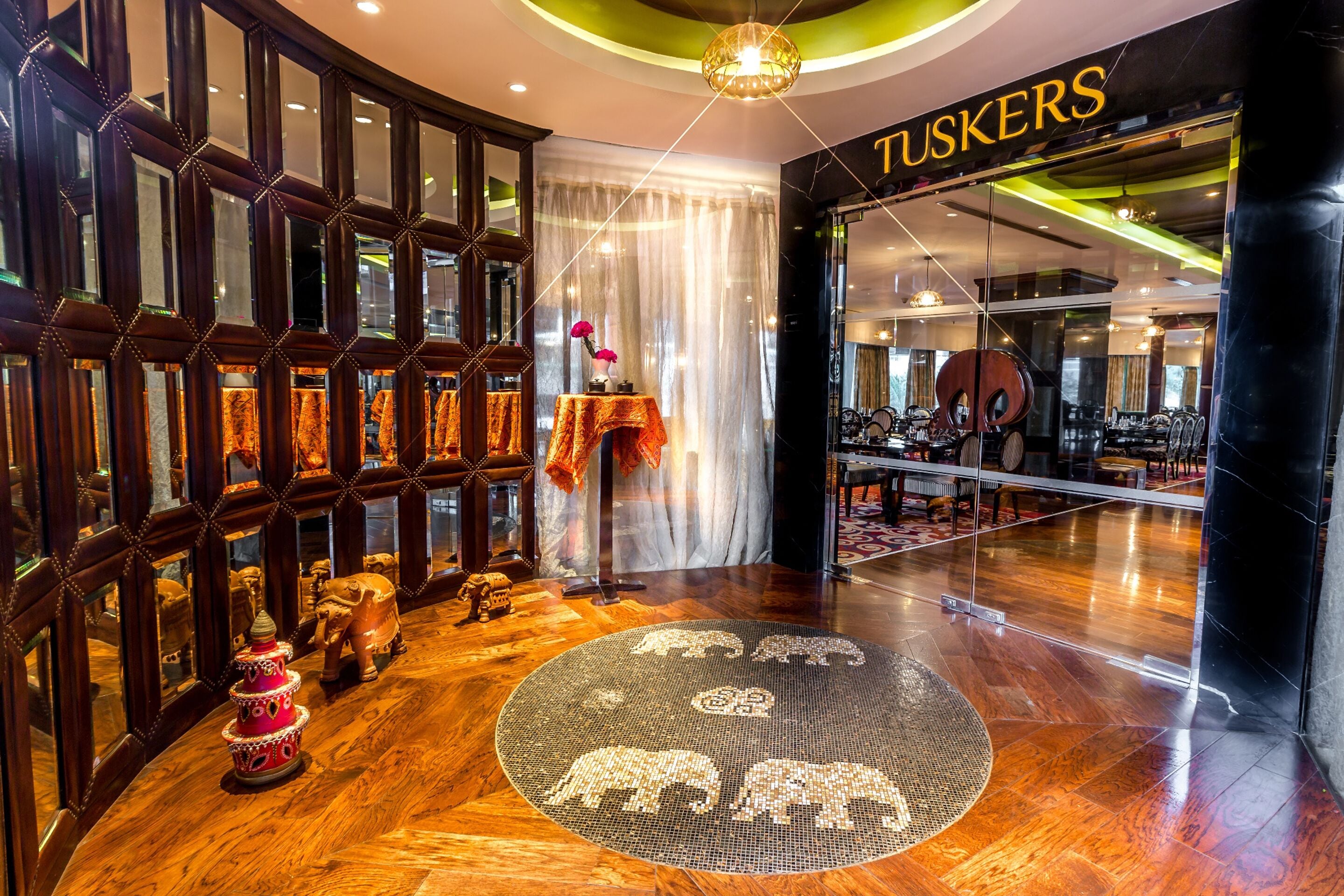 Un vestíbulo de hotel con suelo de madera, un tapete decorativo con diseños de elefantes y una puerta de cristal con el nombre 'TUSKERS'.