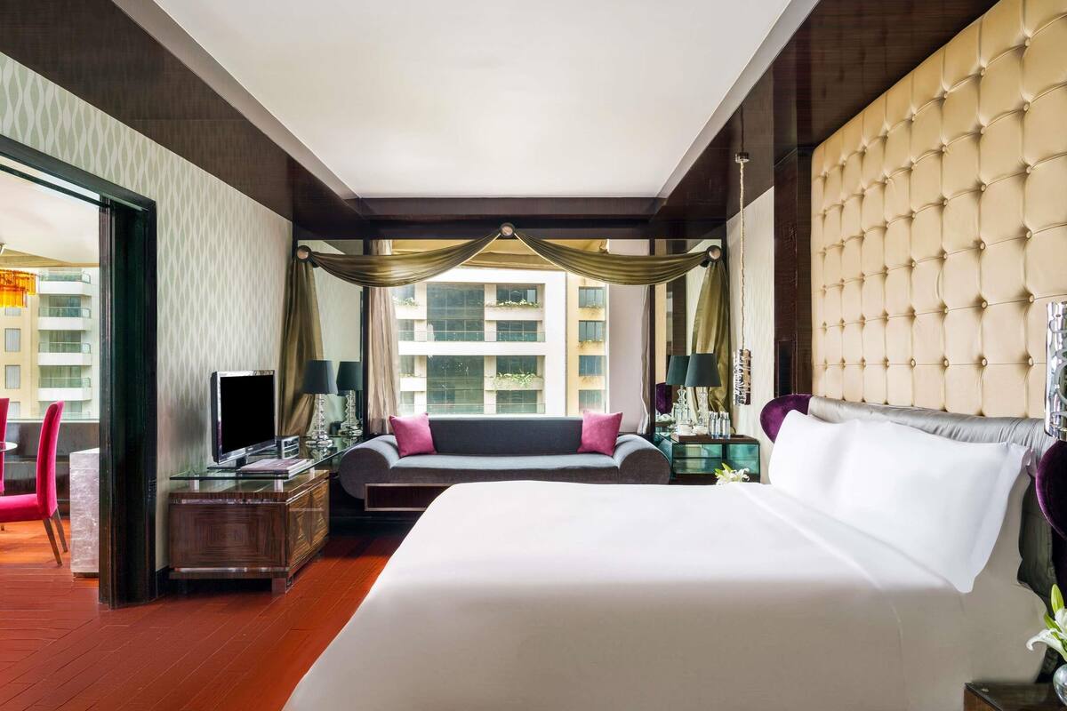 A hotel room with a large bed, a desk, a sofa, and a view of buildings through the window.