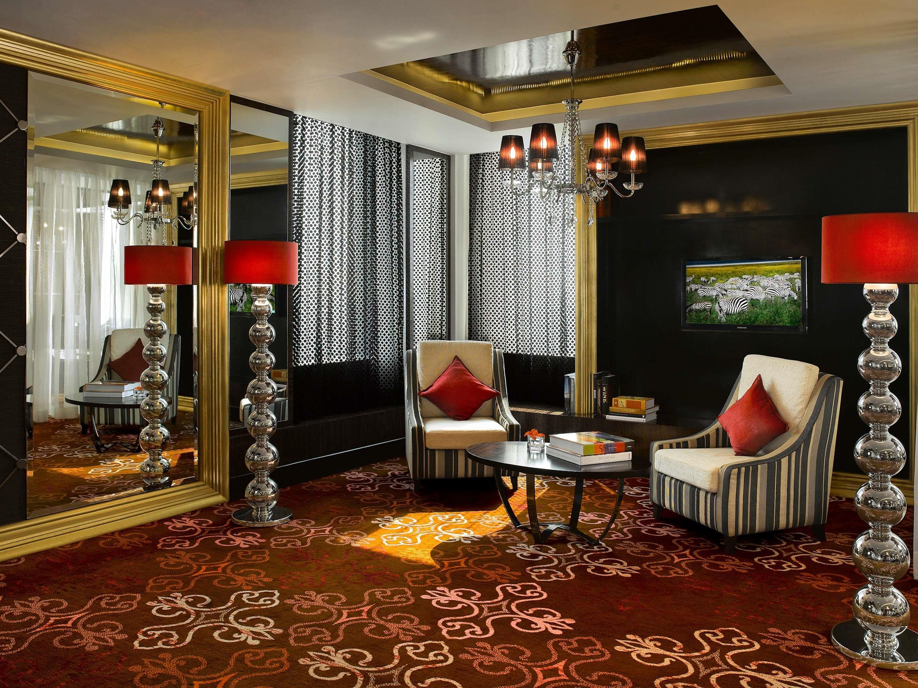 A luxurious room with a large mirror, a chandelier, a red lamp, a striped armchair, and a television on the wall.