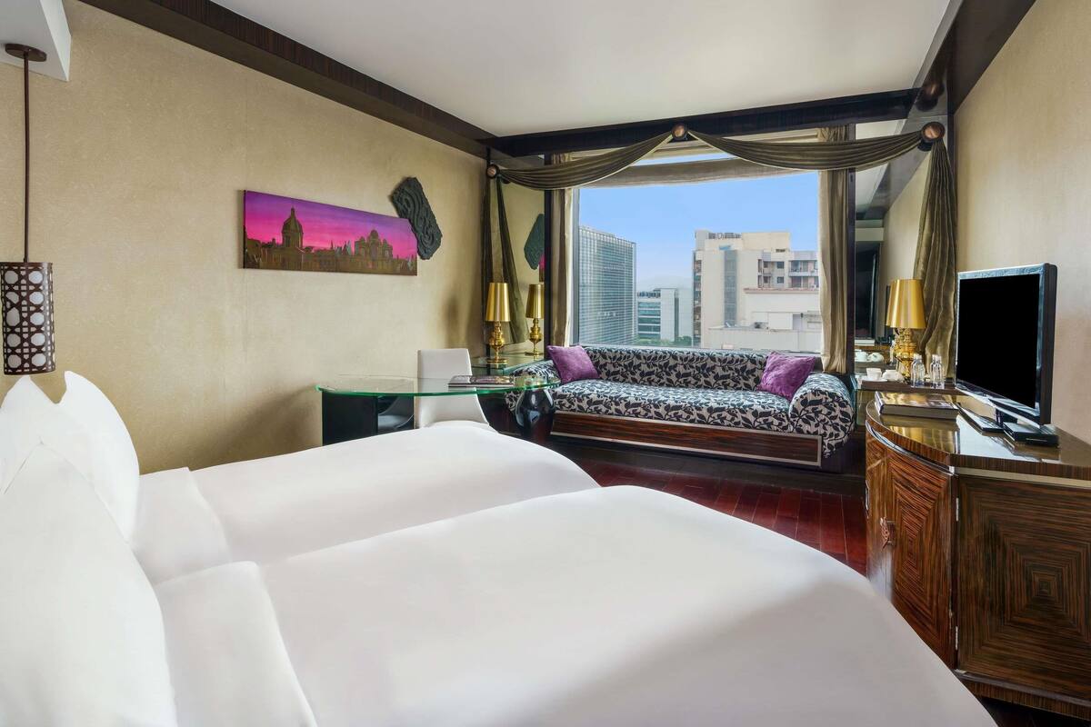 A hotel room with a large bed, a sofa, a desk with a lamp, a TV, and a view of buildings.