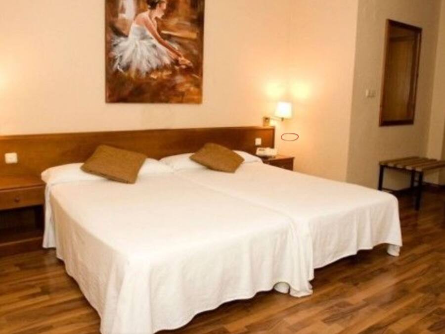 Een tweepersoonsbed met witte lakens en twee kussens, een houten hoofdbord, een nachtkastje met een lamp en een schilderij aan de muur.